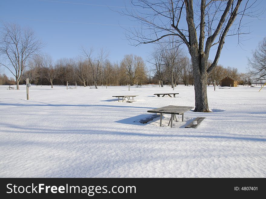 Picnic benches sit idly in the snow on a sunny day waiting for warmer weather to bring the people back. Picnic benches sit idly in the snow on a sunny day waiting for warmer weather to bring the people back.