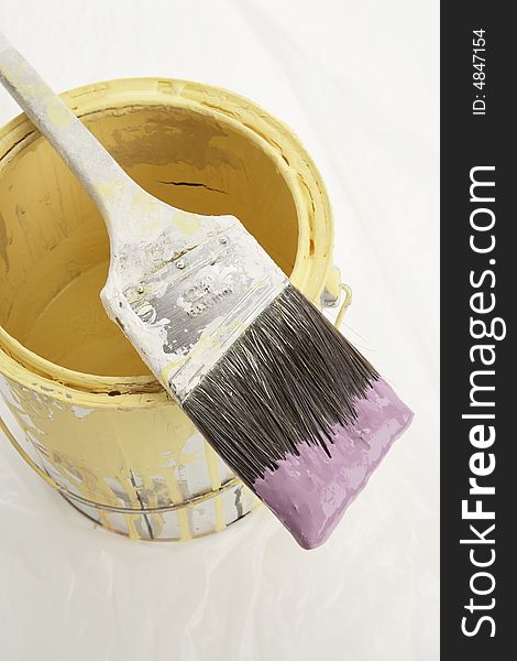 A metallic paint can on top off a plastic drop cloth. The paint can has yellow paint inside. The brush is dipped in purple paint. A metallic paint can on top off a plastic drop cloth. The paint can has yellow paint inside. The brush is dipped in purple paint.