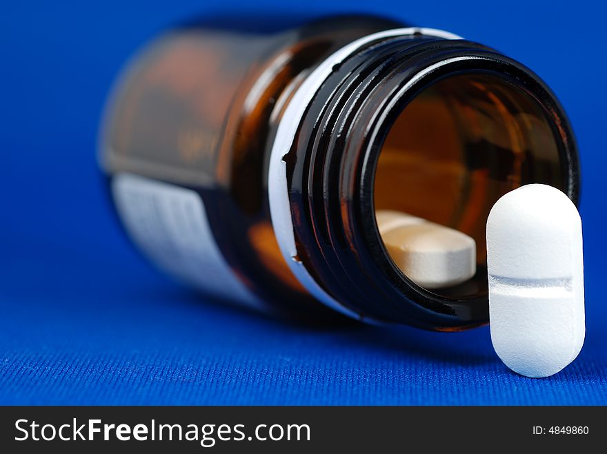 Close up to a pill placed on the right corner of frame together with open bottle of medicine on blue background