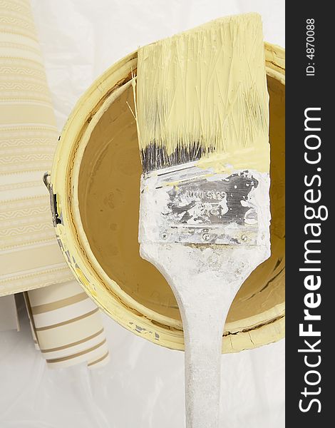 Brush dipped in yellow paint on top of a can of paint with wall paper samples on the side. Brush dipped in yellow paint on top of a can of paint with wall paper samples on the side.