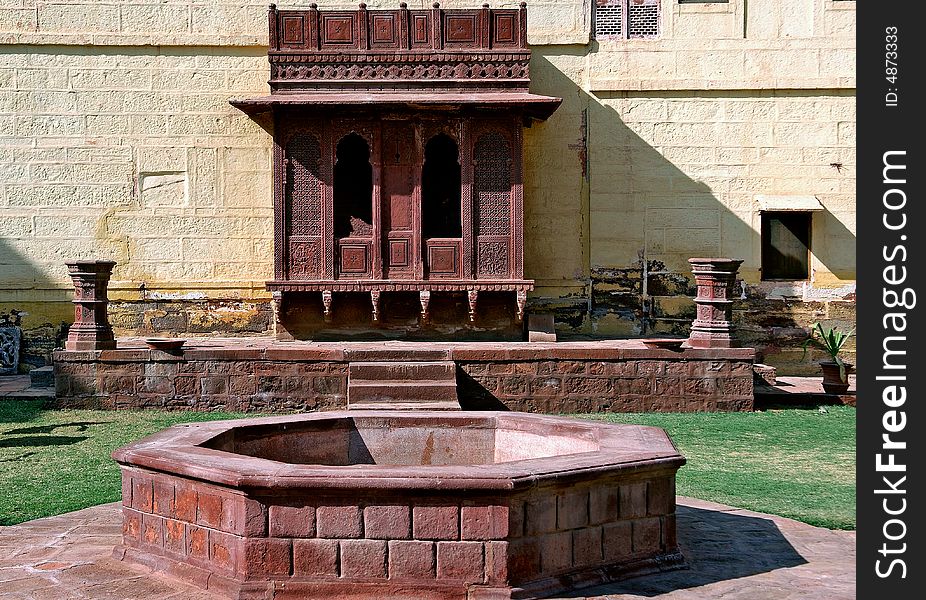 India, Jaisalmer: Indian palace architecture; red walls and a wooden carved frame and window in front of a round stone fountain. India, Jaisalmer: Indian palace architecture; red walls and a wooden carved frame and window in front of a round stone fountain