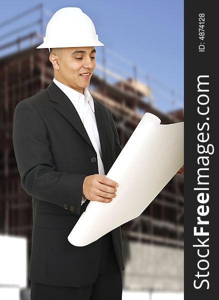 A man wearing hard hat and smiling looking at his floor plan with building under construction behind him. A man wearing hard hat and smiling looking at his floor plan with building under construction behind him
