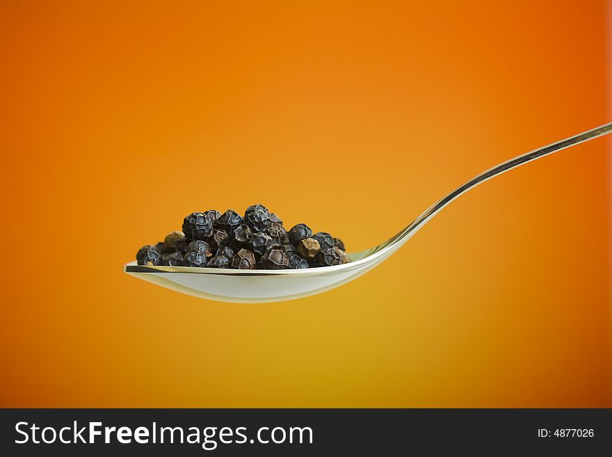 Metal spoon filled with black peppers over orange background