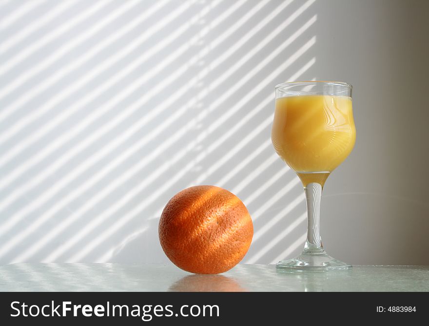 Goblet of orange juice and fresh orange on glass table on background with jalousie shadow. Goblet of orange juice and fresh orange on glass table on background with jalousie shadow