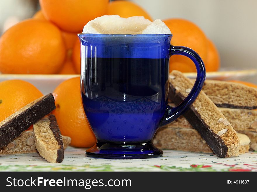 Coffee latte in cobalt blue glass mug surrounded by chocolate biscotti and fresh oranges. Coffee latte in cobalt blue glass mug surrounded by chocolate biscotti and fresh oranges