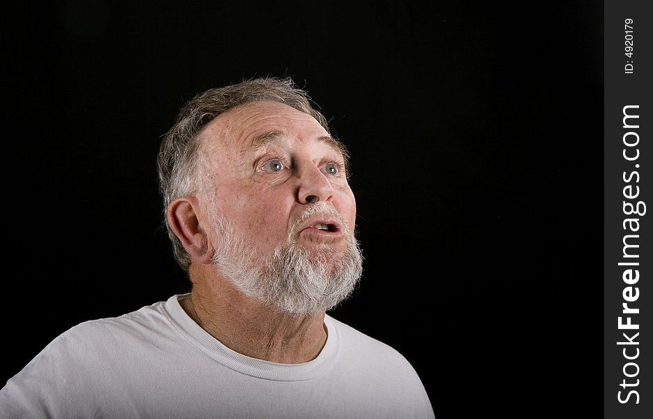 An older man in a white t-shirt on a black background looking up into copy space with shocked expression. An older man in a white t-shirt on a black background looking up into copy space with shocked expression