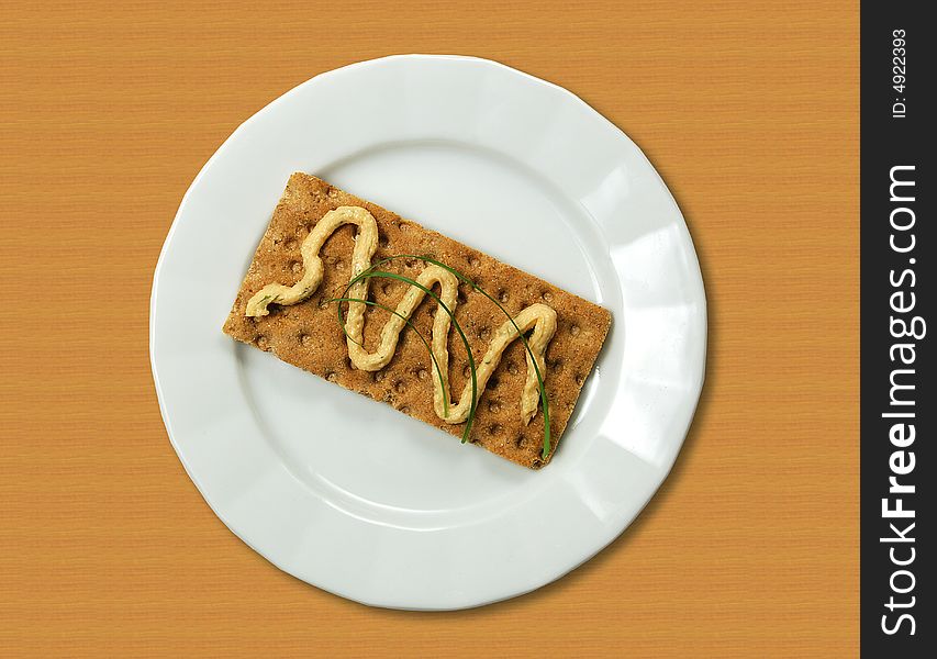A view of a large multi-grained cracker on a plate or saucer and topped with a wavy line of fish spread and green garnish. A view of a large multi-grained cracker on a plate or saucer and topped with a wavy line of fish spread and green garnish.