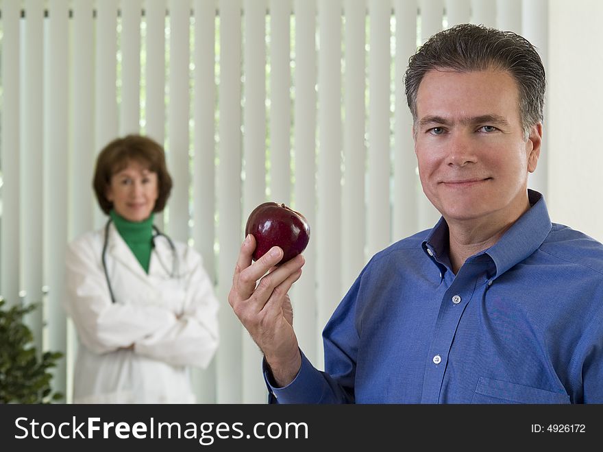 A man with an apple in his hand with a smiling woman dressed as a doctor, out of focus in the background. A man with an apple in his hand with a smiling woman dressed as a doctor, out of focus in the background.