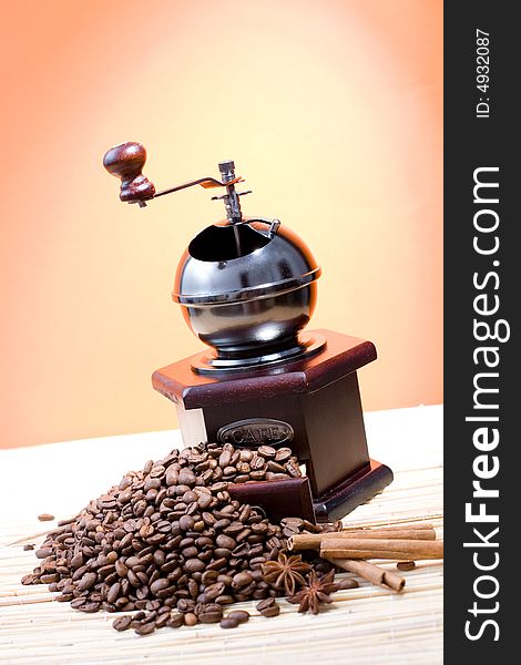 Grinder with coffee beans and sticks of cinnamon on hot orange background.