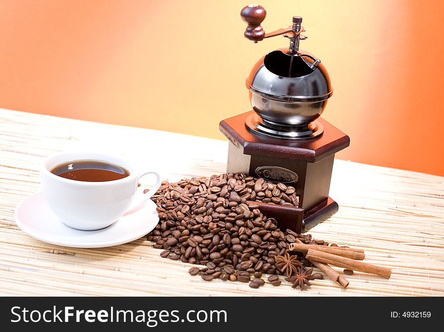 Grinder, cup of coffee, coffee beans and sticks of cinnamon on hot orange background