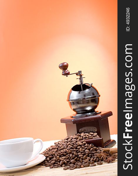 Grinder, cup of coffee, coffee beans and sticks of cinnamon on hot orange background