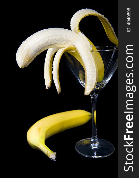 A half peeled banana presented inside a cocktail glass, isolated on a black background.  An unpeeled banana is lying on the side. A half peeled banana presented inside a cocktail glass, isolated on a black background.  An unpeeled banana is lying on the side.