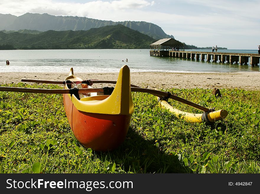 Hawaiian Outrigger Canoe on beach grass with bay, fishing pier and mountains in background.