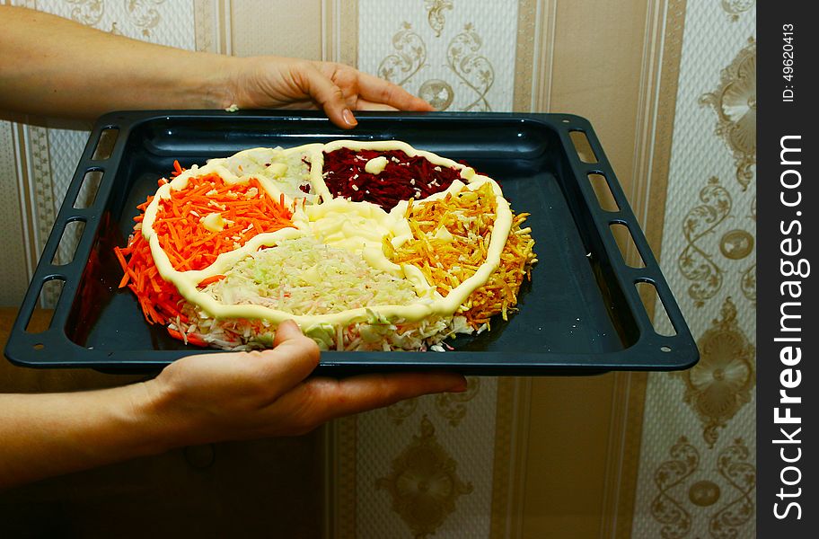 Woman is holding a baking sheet with a salad in the form of cake with carrots, beets, potatoes, onions, mayonnaise