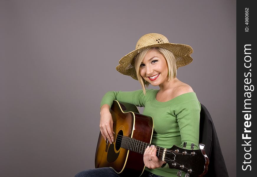 A blonde in a green blouse and straw hat holding a guitar and smiling at the camera. A blonde in a green blouse and straw hat holding a guitar and smiling at the camera