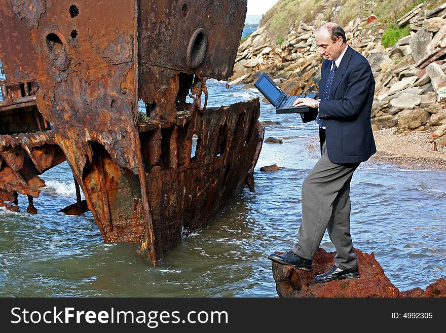 Businessman standing on the rocks at the beach with his laptop and with a beautiful old rusting shipwreck in the background. Businessman standing on the rocks at the beach with his laptop and with a beautiful old rusting shipwreck in the background