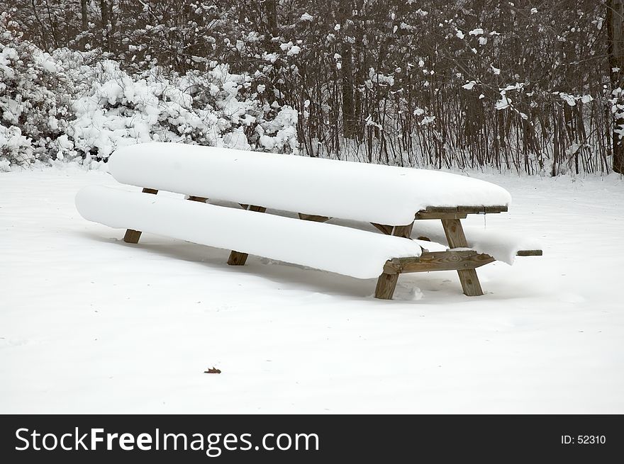 A long picnic table in a park covered by a heavy snowfall. A long picnic table in a park covered by a heavy snowfall.