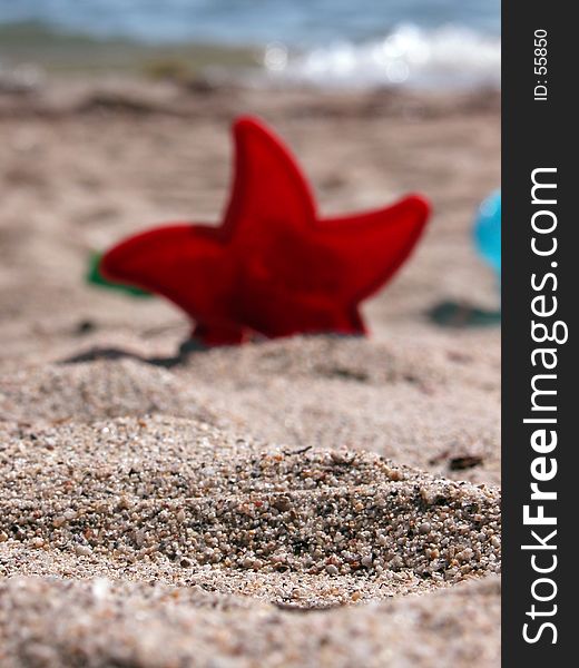 A red plastic star with sea in the background. Sharp sand in the foreground, the rest of the picture is thrown out of focus. A red plastic star with sea in the background. Sharp sand in the foreground, the rest of the picture is thrown out of focus.