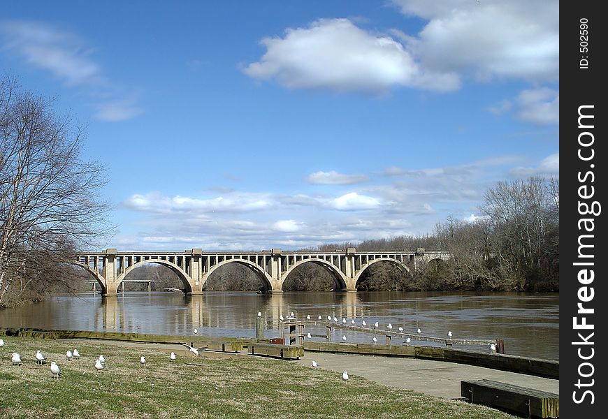 Older railroad bridge lays a nice scene with soft reflections, blue sky with puffy clouds, and seagulls lined up in a row. Older railroad bridge lays a nice scene with soft reflections, blue sky with puffy clouds, and seagulls lined up in a row.