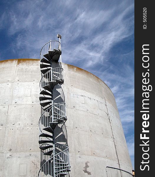 A vast concrete silo with metal staircase on the side beneath wispy clouds in a blue sky. A vast concrete silo with metal staircase on the side beneath wispy clouds in a blue sky