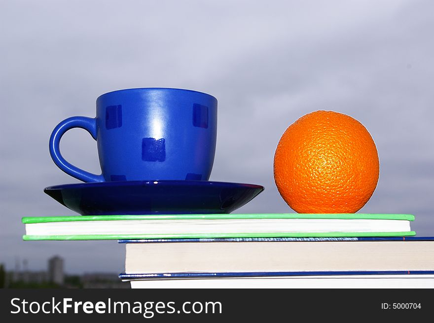 Orange, cup and books on a background of the sky. Orange, cup and books on a background of the sky