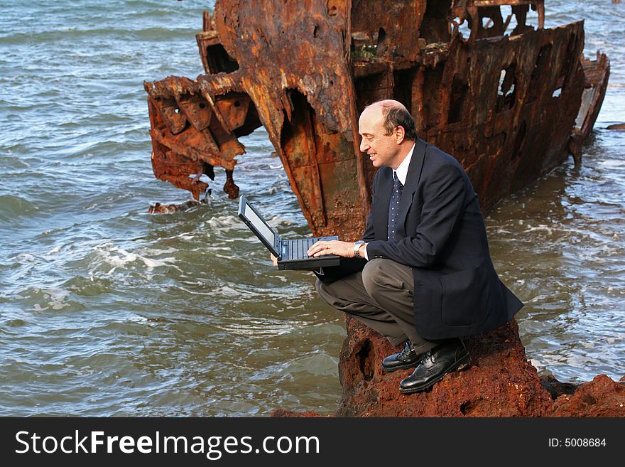 Businessman sitting on the rocks at the beach with his laptop and with a beautiful old rusting shipwreck in the background. Businessman sitting on the rocks at the beach with his laptop and with a beautiful old rusting shipwreck in the background