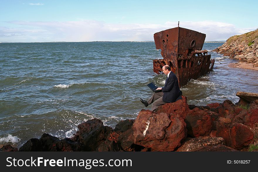 Businessman sitting on the rocks at the beach with his laptop and with a beautiful old rusting shipwreck in the background. Businessman sitting on the rocks at the beach with his laptop and with a beautiful old rusting shipwreck in the background