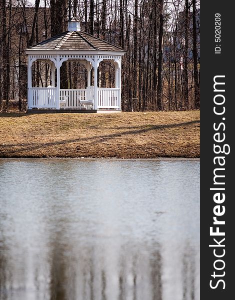 A white gazebo between a pond  and a stand of leafless trees. A white gazebo between a pond  and a stand of leafless trees.