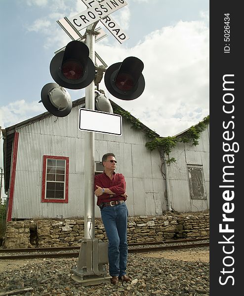 A man wearing dark sunglasses, leaning up against a railroad crossing signal lights pole.