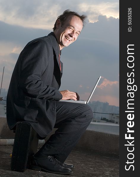 A business man, sitting on his briefcase, working on his laptop outdoors, while smiling. A business man, sitting on his briefcase, working on his laptop outdoors, while smiling.