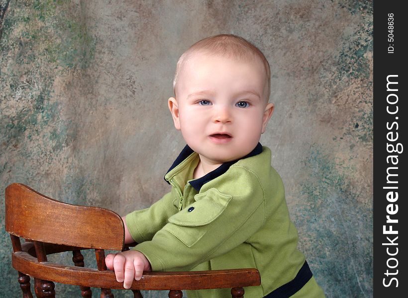 Baby boy standing in front of muslin background beside old wooden chair. Baby boy standing in front of muslin background beside old wooden chair