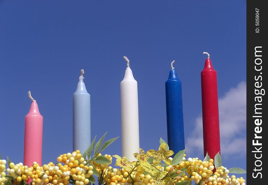A set of five colorful candles are standing in between some flowers, against the blue sky. A set of five colorful candles are standing in between some flowers, against the blue sky