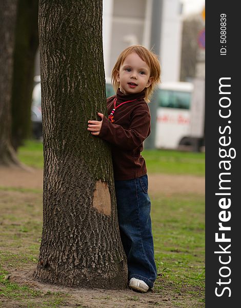 Little beauty girl playing hide-and-seek at the park. She hideing round the big old tree with split off part of bark. Little beauty girl playing hide-and-seek at the park. She hideing round the big old tree with split off part of bark.