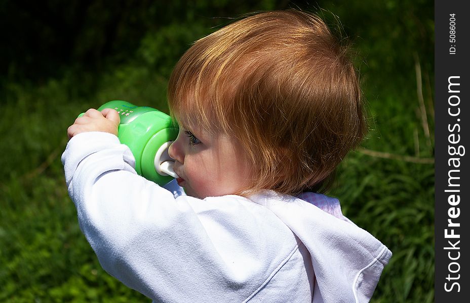 The little girl drinks from a green bottle on a background of a grass