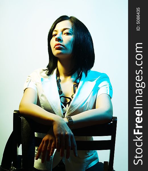 A high-key portrait about an attractive trendy girl with black hair who is lighted with blue and yellow, she is sitting on a chair and she has a charming look. She is wearing black pants, a white coat and a stylish necklace. A high-key portrait about an attractive trendy girl with black hair who is lighted with blue and yellow, she is sitting on a chair and she has a charming look. She is wearing black pants, a white coat and a stylish necklace.