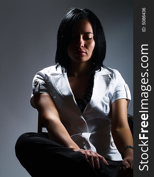 A low-key portrait about an attractive trendy girl with black hair who is sitting on a chair and she is thinking. She is wearing black pants and a white stylish coat. A low-key portrait about an attractive trendy girl with black hair who is sitting on a chair and she is thinking. She is wearing black pants and a white stylish coat.