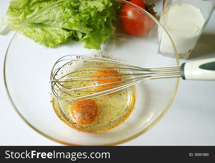 Two cracked eggs and a whisk in a bowl surrounded by a glass of milk, tomato and salad. Two cracked eggs and a whisk in a bowl surrounded by a glass of milk, tomato and salad