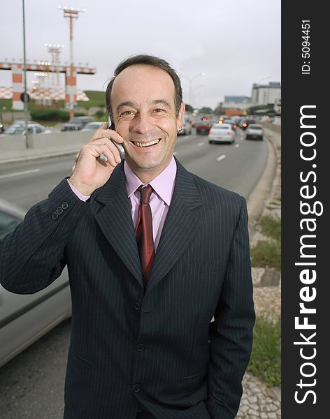 Latin american businessman standing by a busy highway talking on his cell phone and smiling. Latin american businessman standing by a busy highway talking on his cell phone and smiling