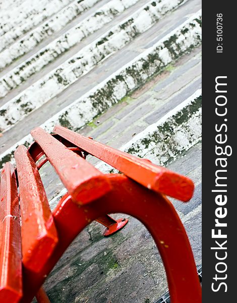 Close up of a red wooden seat with, on the back, old white stairs made of stones. This can be a concept image with the implication of a choice between stay and go. Useful also for relax and rest concept. Focus on the center of the seat. Close up of a red wooden seat with, on the back, old white stairs made of stones. This can be a concept image with the implication of a choice between stay and go. Useful also for relax and rest concept. Focus on the center of the seat.