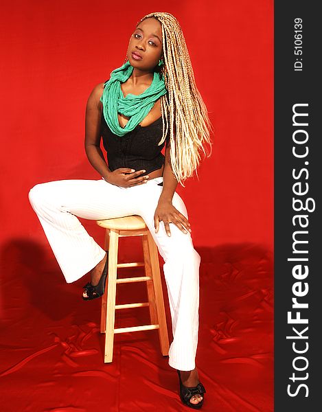 An friendly blond woman in white pants and black top and green scarf sitting 
in an studio on a bar chair for red background. An friendly blond woman in white pants and black top and green scarf sitting 
in an studio on a bar chair for red background.