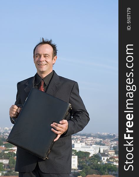 Businessman standing on a city rooftop and smiling while holding his briefcase close to his chest. Businessman standing on a city rooftop and smiling while holding his briefcase close to his chest