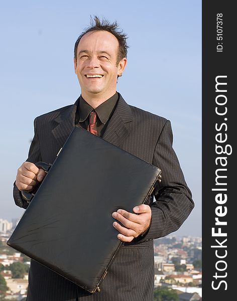 Businessman standing on a city rooftop and smiling while holding his briefcase close to his chest. Businessman standing on a city rooftop and smiling while holding his briefcase close to his chest