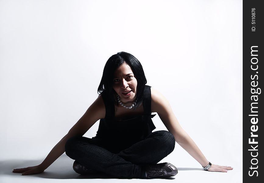 A portrait about an attractive trendy girl with black hair who is sitting, she is spreading her arms on ground and she is smiling. She is wearing a black dress, blue jeans and a white necklace. A portrait about an attractive trendy girl with black hair who is sitting, she is spreading her arms on ground and she is smiling. She is wearing a black dress, blue jeans and a white necklace.