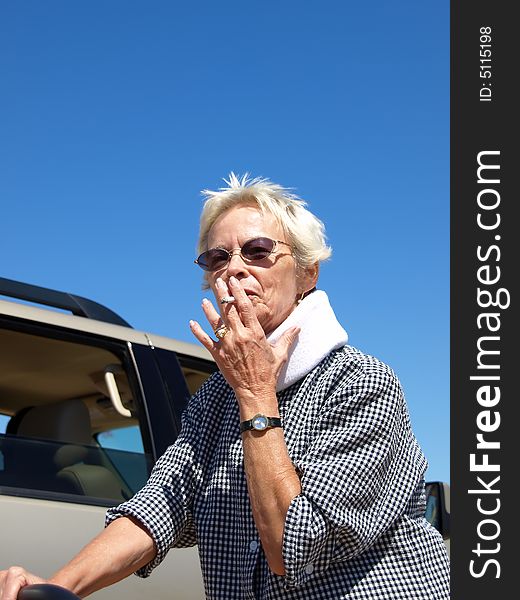 Older woman with close-cropped white hair taking a drag from a cigarette while standing outside in the desert. Shot is framed against a clear blue sky and desert sand. Older woman with close-cropped white hair taking a drag from a cigarette while standing outside in the desert. Shot is framed against a clear blue sky and desert sand.