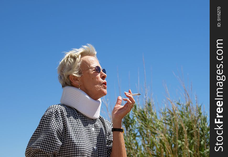 Older woman with close-cropped white hair taking a drag from a cigarette while standing outside in the desert. Shot is framed against a clear blue sky and desert sand. Older woman with close-cropped white hair taking a drag from a cigarette while standing outside in the desert. Shot is framed against a clear blue sky and desert sand.