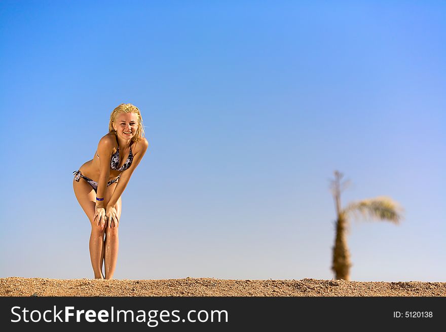Young woman standing on sand leaning on her knees and waiting for something. Blue sky and palm on a background. there is a good place for your text