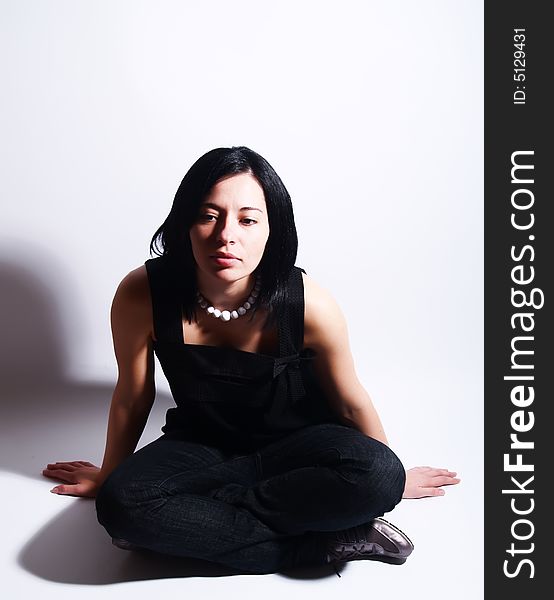 A high-key portrait about an attractive trendy girl with black hair who is sitting, she is spreading her arms on ground and she is thinking. She is wearing a black dress, blue jeans and a white necklace. A high-key portrait about an attractive trendy girl with black hair who is sitting, she is spreading her arms on ground and she is thinking. She is wearing a black dress, blue jeans and a white necklace.