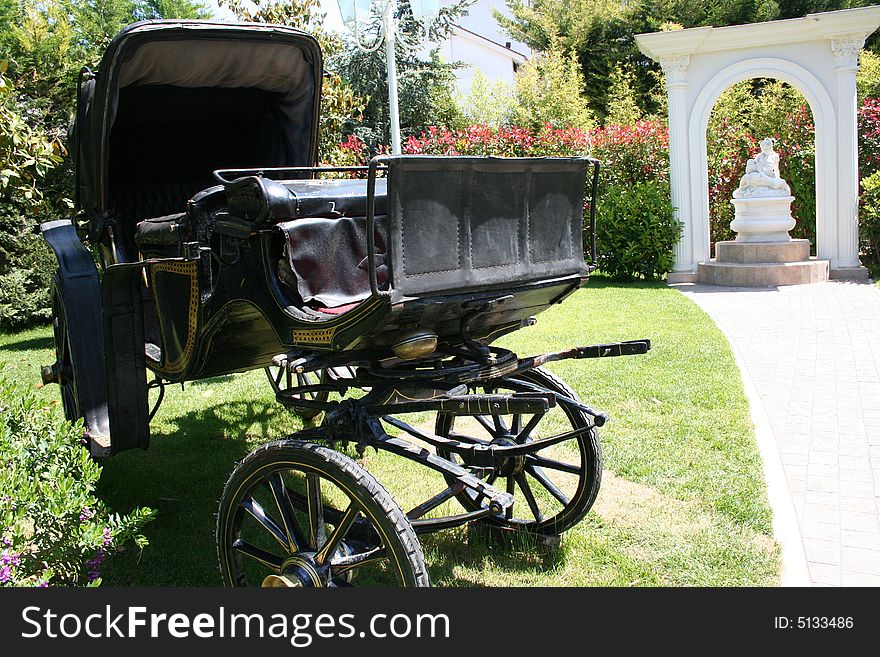 There is an old black coach parked in a park. There are Trees, grass and a statue on the background.