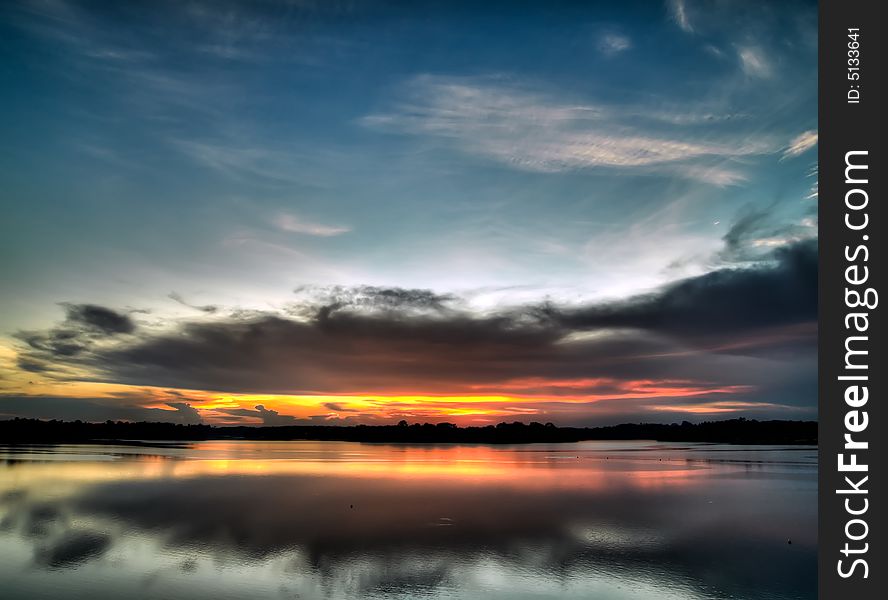 Sun setting below low altitude clouds while illuminating high altitude ones over a still lake. Sun setting below low altitude clouds while illuminating high altitude ones over a still lake