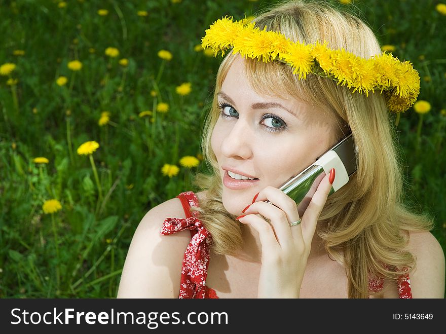 Young beautiful blond hair woman in garland talking on the mobile phone, sitting on the grass among dandelions. Young beautiful blond hair woman in garland talking on the mobile phone, sitting on the grass among dandelions